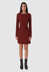 Abito lungo a maniche lunghe color marrone scuro con design frontale intrecciato. Tessuto liscio. Abbinato a stivaletti neri con borchie alla caviglia. Semplice scollo tondo.