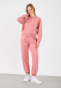 Pulover roz deschis și pantaloni de trening asortați, confecționați dintr-un material moale, având o potrivire lejeră și manșete elasticizate, complementar cu adidași albi.