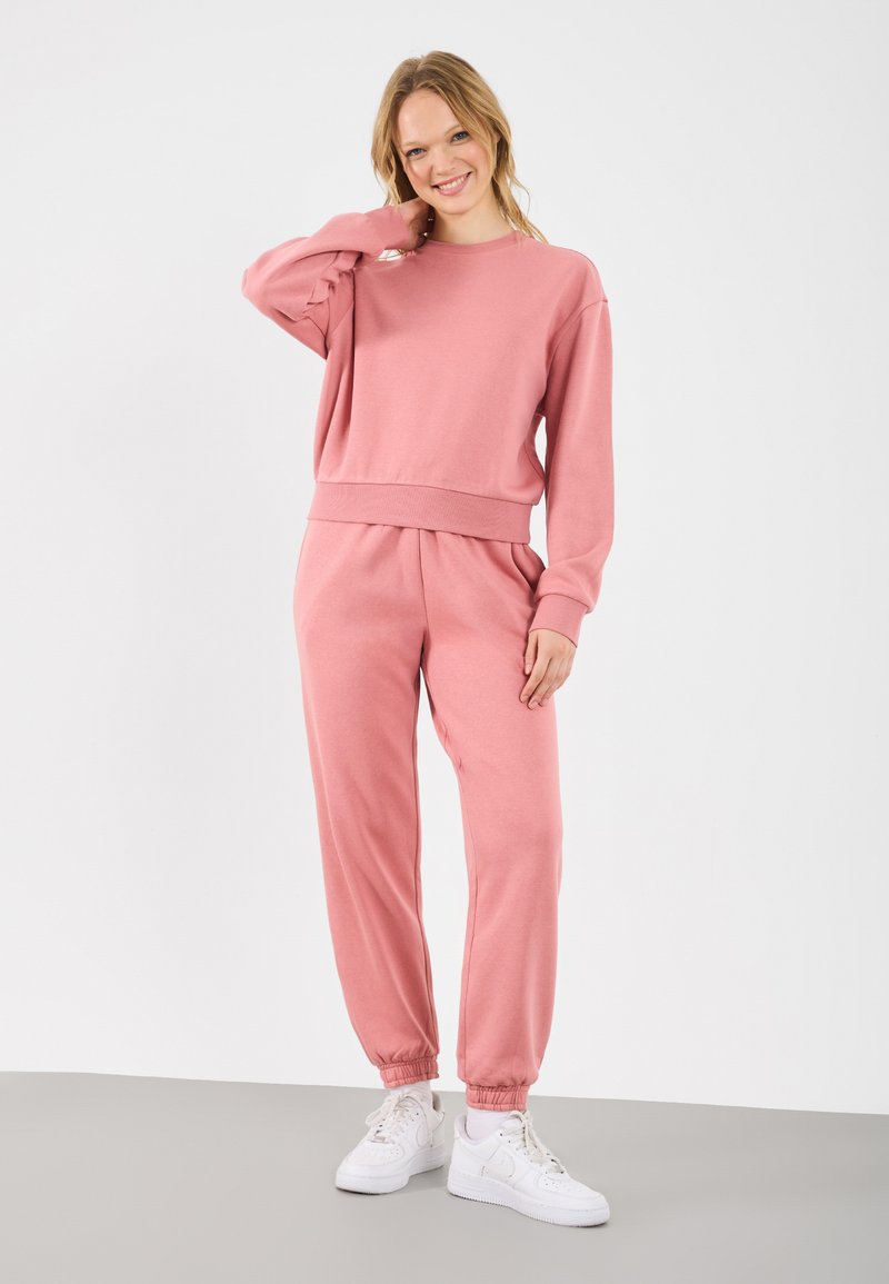 Pulover roz deschis și pantaloni de trening asortați, confecționați dintr-un material moale, având o potrivire lejeră și manșete elasticizate, complementar cu adidași albi.