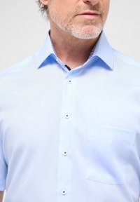 Lichtblauw shirt met korte mouwen, met een knoopsluiting aan de voorkant, een borstzak en een patroon aan de binnenkraag. De stof lijkt glad en lichtgewicht.