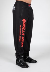 Sorte træningsbukser med et struktureret stof, udstyret med iøjnefaldende røde "GORILLA WEAR" bogstaver og en lynlåslomme på siden.