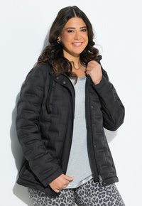 Černá prošívaná péřová bunda s kapucí, texturovaná tkanina, přední zip a stahovací šňůrka. Nositá přes světle šedou košili s metalickým okrajem.
