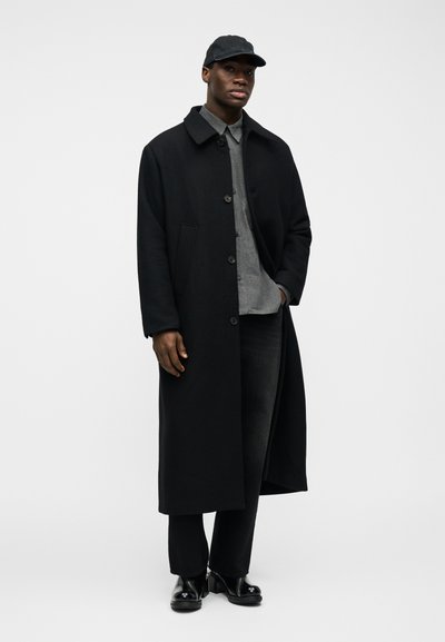 Μαύρος άνδρας που φοράει ένα μακρύ μαύρο παλτό πάνω από γκρι πουκάμισο με κουμπιά, μαύρο παντελόνι, μαύρο καπέλο και μαύρα στιλβωμένα παπούτσια, στέκεται μπροστά σε λευκό φόντο.