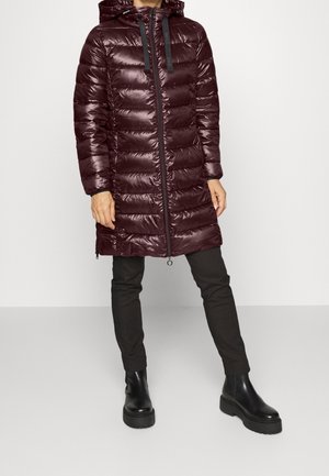 Personne portant un long manteau matelassé brillant couleur bordeaux foncé avec capuche, pantalon noir et bottines noires épaisses, debout devant un fond uni.