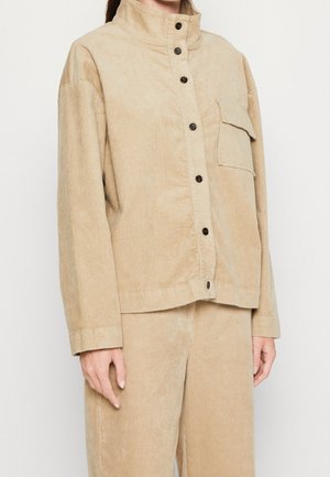 Beige kordfløjlsjakke med sorte knapper og en klaplomme på brystet, båret sammen med matchende kordfløjlbukser.