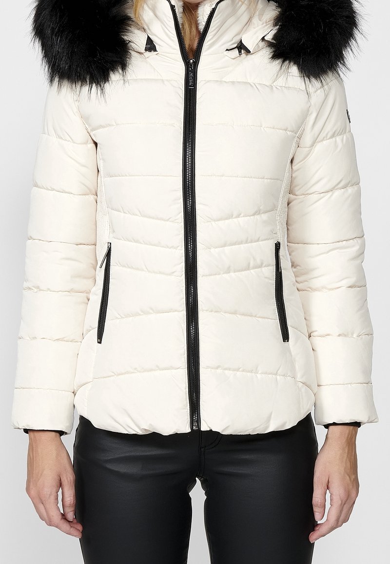 Giacca puffa di un crema chiaro con zip e tasche nere, dotata di cappuccio in pelliccia nera removibile e texture trapuntata.