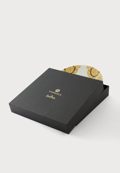 Μαύρο τετράγωνο κουτί Versace με χρυσό λογότυπο, μερικώς ανοιχτό, αποκαλύπτοντας μια περίτεχνη κίτρινη και λευκή διακοσμητική πιατέλα μέσα.
