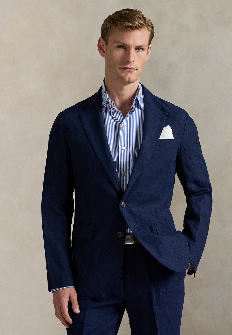 Navy linen blazer with two buttons and flap pockets, paired with a light blue and white striped shirt. White pocket square adds detail.