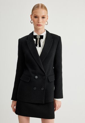Blazer negro de doble botonadura con tejido texturizado, grandes botones y bolsillos frontales. Llevado sobre una camisa blanca con una pajarita decorativa.