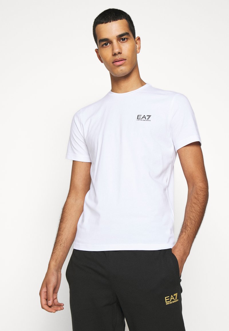 Camiseta blanca de algodón con cuello redondo y mangas cortas, con un pequeño logo negro de EA7 en el lado izquierdo del pecho.