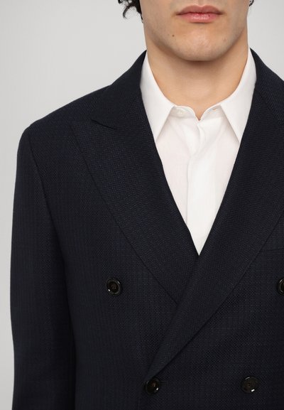 Blazer texturat de culoare bleumarin, cu două rânduri de nasturi, rever înclinat, nasturi negri și un guler de cămașă alb vizibil dedesubt.