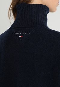 Marinblå fleecetröja med en hög ribbad krage. Har en märkesetikett på baksidan med vita, röda och blå accenter.