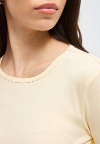 Kobieta o długich ciemnych włosach, ubrana w jasnżółtą koszulkę z okrągłym dekoltem, pokazana od szyi do barku, zwrócona lekko w lewo.