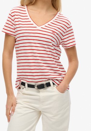 Femme portant un T-shirt à col en V rayé blanc et rouge et un pantalon blanc avec une ceinture noire, la main dans la poche, devant un fond uni.