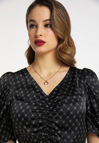 Schwarze Bluse mit satinartigem Finish, Puffärmel-Design, gerafftem Vorderteil und sich wiederholendem weißen Muster. Goldkette mit Anhänger.