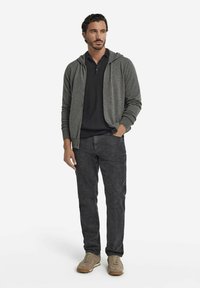 Man står klädd i en mörkgrå zip-up hoodie, svart polo skjorta, mörka byxor och beige sneakers, med en hand i fickan.