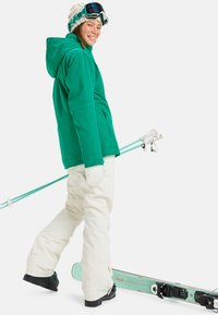 Groene waterdichte jas met een capuchon, crème skibroeken, witte handschoenen en groene skistokken. Skiuitrusting klaar voor wintersport.