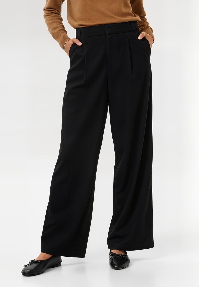 Pantalons larges noirs en tissu lisse, dotés de plis sur le devant et de poches latérales. Associés à des ballerines noires simples.