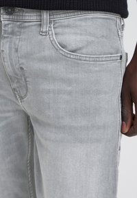 Nærbillede af lysegrå jeans med frontlomme og sort strikket top, viser en del af en mørkhudet persons hånd ved siden af.