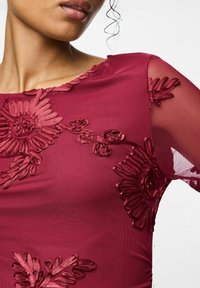 Bordó šaty s průsvitnými síťovanými rukávy, zdobené vyvýšenou květinovou výšivkou v tónovaném designu. Detailní záběr na výstřih a ramenní detaily.