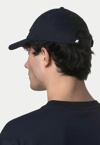 Homme aux cheveux courts et foncés portant une casquette de baseball noire et une chemise noire, vu de dos et légèrement tourné vers la gauche.