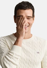 Baltas megztinis su pynimo raštu, ribinėmis rankogaliais ir mažu logotipu ant krūtinės. Modelis šypsosi, viena ranka dangstydamas veidą.