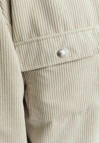 Light beige corduroy fabric with vertical ribbing. Features a large pocket with a silver snap button accent. Soft texture with visible lines.