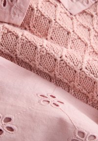 Lyserødt stof med et struktureret strikket mønster og broderede blomsterdetaljer. Materialerne inkluderer en blød strik og let bomuld.