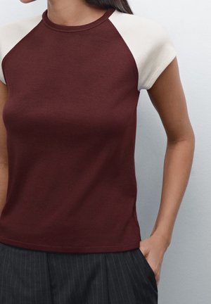 Femme portant un haut bordeaux à manches courtes avec des manches blanches et un pantalon noir à fines rayures, une main dans la poche, devant un fond uni.