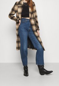 Abrigo de cuadros en tonos marrón y beige, combinado con jeans de cintura alta azules y botines negros. Mangas largas, con botones en la parte delantera.