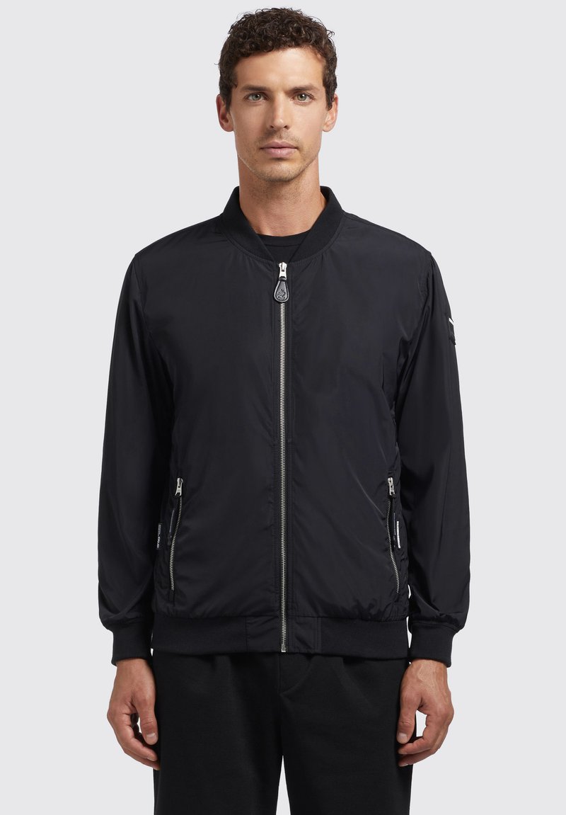 Jachetă bomber neagră cu fermoar frontal, buzunare laterale, manșete și guler ribbed, realizată dintr-un material ușor, neted, cu un design minimalist.