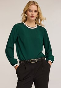 Blusa verde a maniche lunghe con colletto girocollo in bianco e nero, tessuto texture e vestibilità morbida, abbinata a pantaloni neri a vita alta e una cintura con borchie.