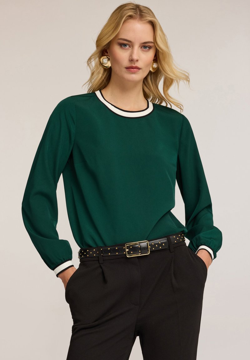 Blusa verde a maniche lunghe con colletto girocollo in bianco e nero, tessuto texture e vestibilità morbida, abbinata a pantaloni neri a vita alta e una cintura con borchie.