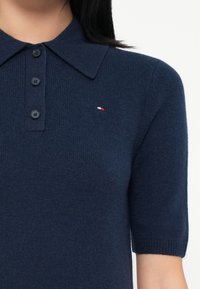 Polo majica kratkih rukava tamnoplave boje s ovratnikom i tri dugmeta, s malim logotipom u crvenoj, bijeloj i tamnoplavoj boji na prsima.