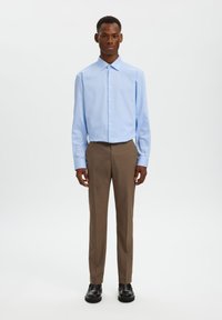 Ανοιχτό μπλε πουκάμισο με κουμπιά και ανοιχτό γιακά, συνδυασμένο με καφέ παντελόνια και μαύρα παπούτσια. Το σύνολο χαρακτηρίζεται από απλά σχεδιαστικά στοιχεία.