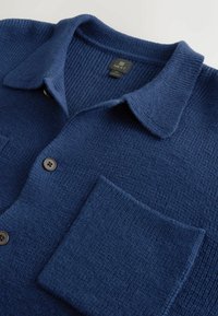 Granatowy kardigan wykonany z fakturowanej dzianiny z kołnierzykiem, z dużą przednią kieszenią i czarnymi guzikami. Widoczna metka marki.