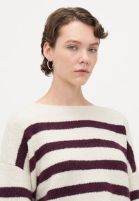 Junge Frau mit kurzen, lockigen Haaren trägt silberne Creolen und einen weißen Pullover mit dunkelvioletten horizontalen Streifen.