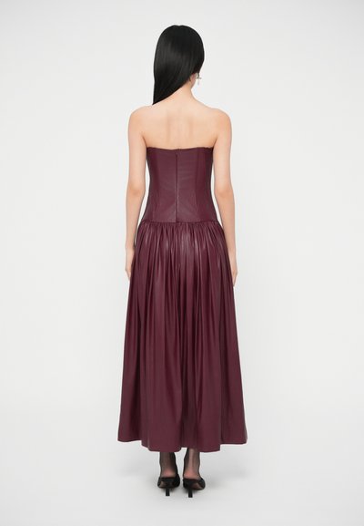 Bordové kožené šaty bez ramienok s priliehavým živôtikom, nariasenou dlhú sukňou a zapínaním na zips vzadu. Rovné záhyby vytvárajú plynulú siluetu.