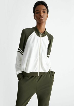 Giacca bomber verde oliva e bianca con zip, maniche raglan, tasche frontali e tre strisce bianche su ciascuna manica. Texture liscia.