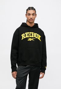 Čierna mikina s kapucňou so veľkým žltým logom "Reebok" a grafickým dizajnom nižšie. Obsahuje predné vrecko a rebrované manžety. Nosená s čiernymi nohavicami.