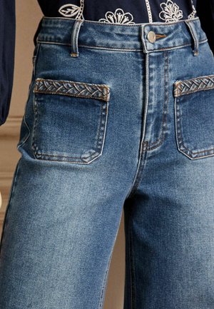 Jean en denim bleu taille haute avec des accents tressés sur les poches arrière, présentant un design classique à cinq poches et des jambes légèrement fuselées.