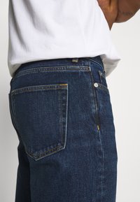 Tmavé džíny z denimu s viditelným šitím, obsahující standardní zadní kapsu a malý kovový knoflík a zapínání. Tkanina má hladkou strukturu.