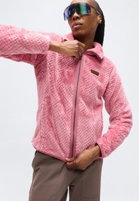 Άτομο που φοράει μεγάλα αντανακλαστικά γυαλιά ηλίου και ένα ροζ υφασμάτινο μπουφάν με φερμουάρ, τραβώντας το φερμουάρ προς τα πάνω με το ένα χέρι.