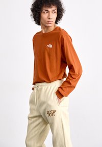 Μακρυμάνικη πορτοκαλί μπλούζα της The North Face; μπεζ παντελόνια φόρμας με ρίγες και γραφικό "Just Do It" σε καφέ.