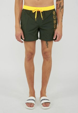 Uomo con pantaloncini da bagno verde scuro con fascia gialla in vita e coulisse, ciabatte bianche e tatuaggi visibili sulle gambe, in piedi su sfondo semplice.