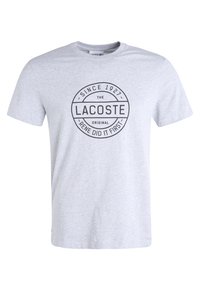 Graues Baumwoll-T-Shirt mit kurzen Ärmeln, das ein kreisförmiges schwarzes Grafikdesign mit den Texten "LACOSTE", "Seit 1927" und "Rene Did It First" zeigt.