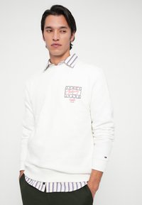 Mand med mørkt hår iført en hvid Tommy Jeans sweatshirt over en stribet skjorte, hænderne i lommerne, ser til siden mod en ensfarvet baggrund.