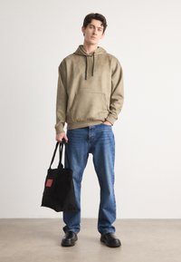 Um hoodie verde-oliva com um bolso frontal, combinado com calças de ganga azuis. O modelo segura uma bolsa tote preta com um rótulo vermelho, usando sapatos pretos.