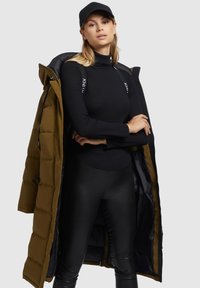 Long olive green puffer coat, unzipped, revealing a fitted black turtleneck and shiny black leggings. Black baseball cap completes the look.