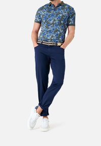 Kortärmad polo t-shirt i mörk olivgrön med blå palmblads-mönster, tillsammans med marinblå byxor och vita sneakers, med händerna i fickorna.